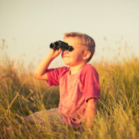 A young boy is looking through binoculars while hiding in the tall grass of an English field. He could be spying on or searching for something newsworthy or just bird watching. Image taken in the sunset light and with shallow depth of field.